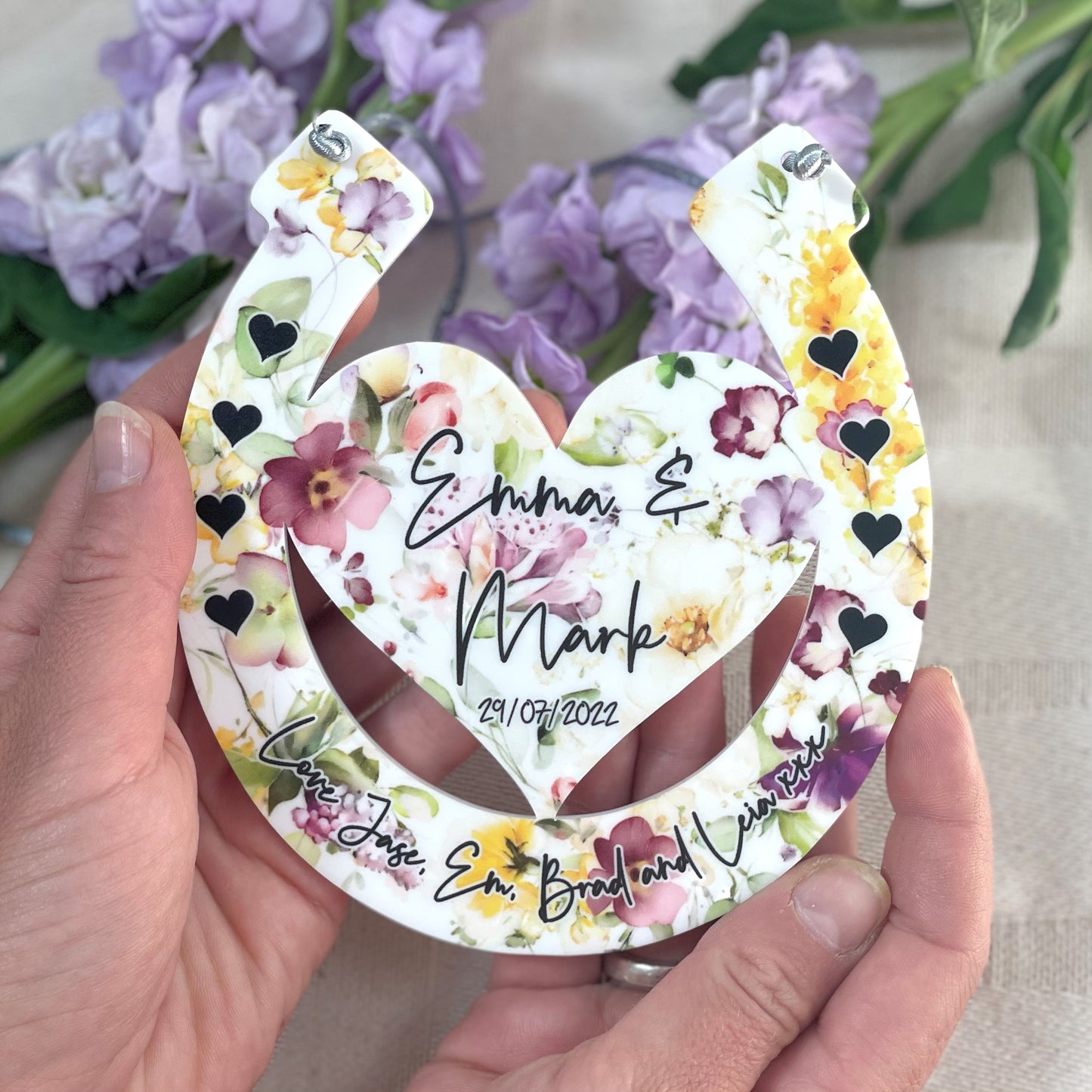 Floral horseshoe design with engraved names being held up by hands with a floral background front shot