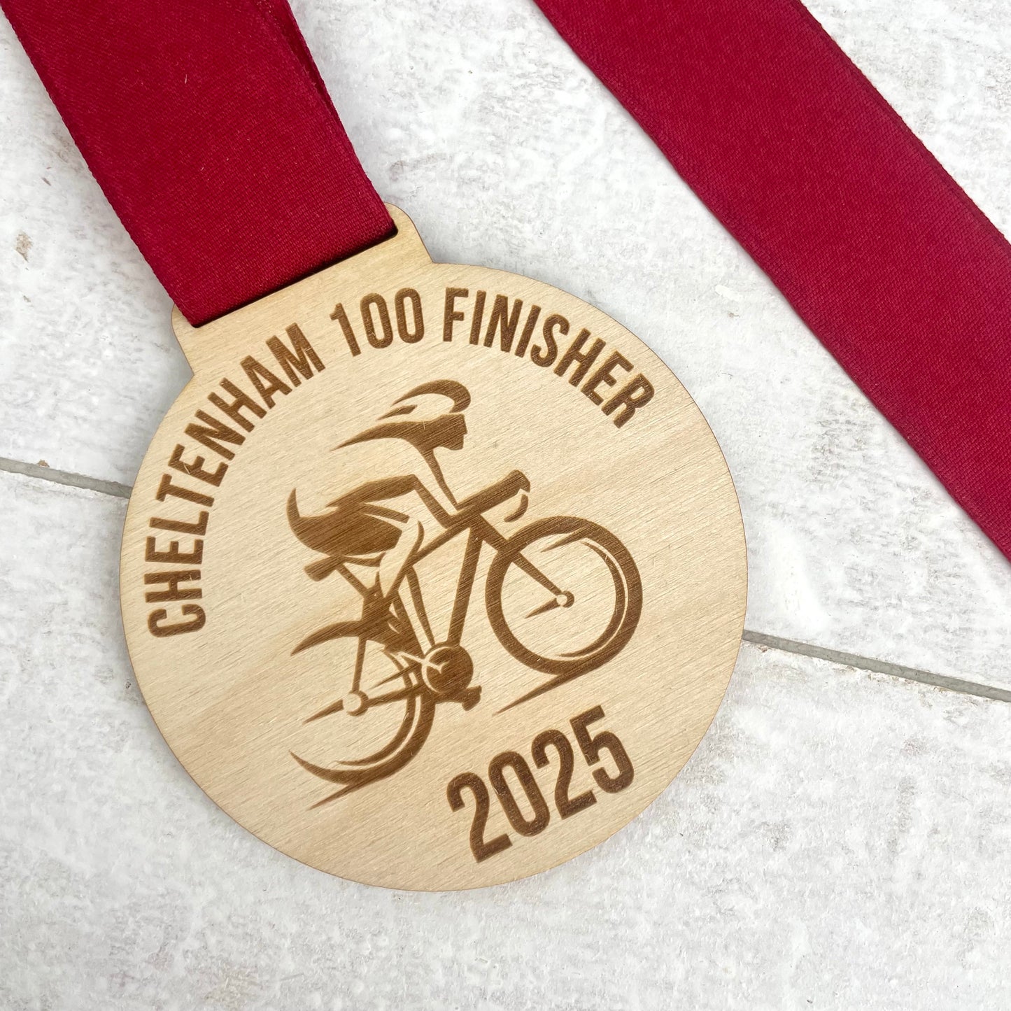 Eco-friendly wooden medal with 'Cheltenham 100 Finisher 2025' text and cyclist with a maroon ribbon.