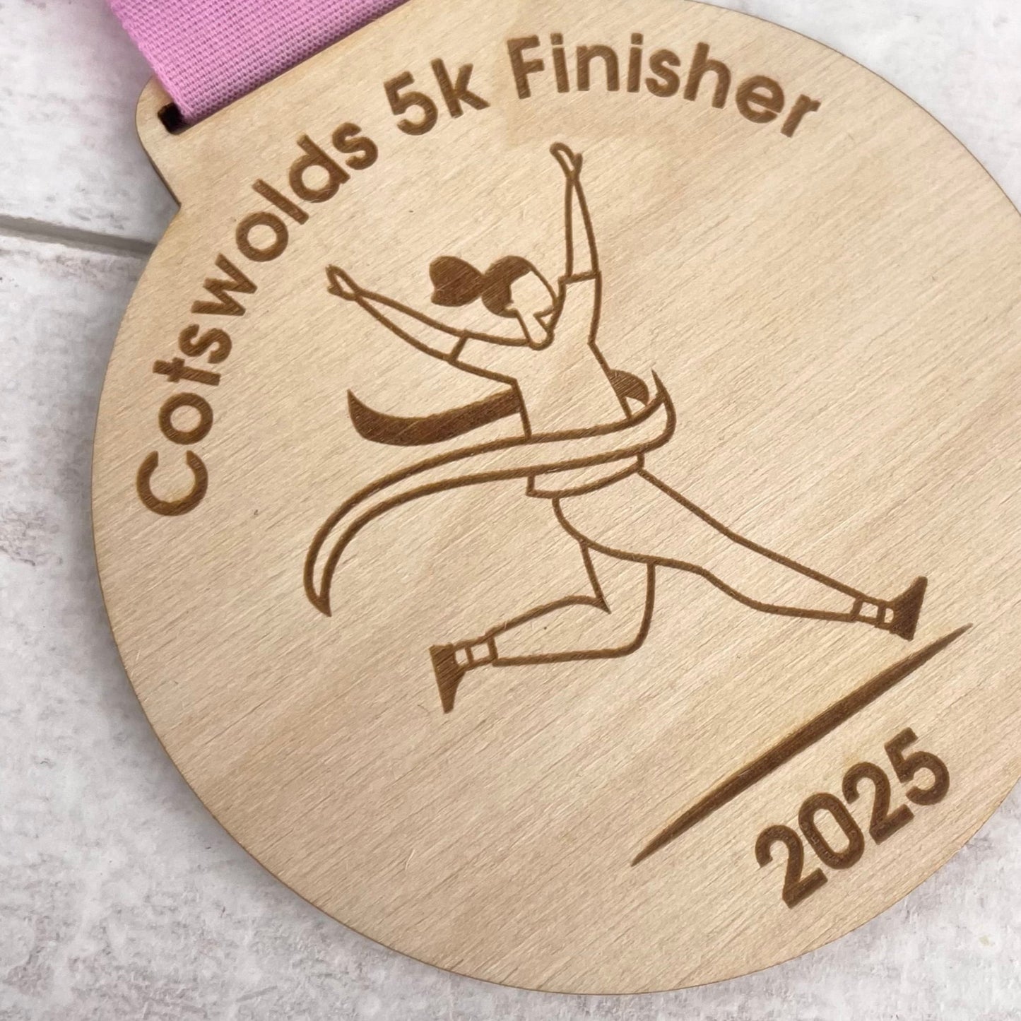Wooden medal with 'Cotswolds 5k Finisher' '2025' text and a running person illustration.