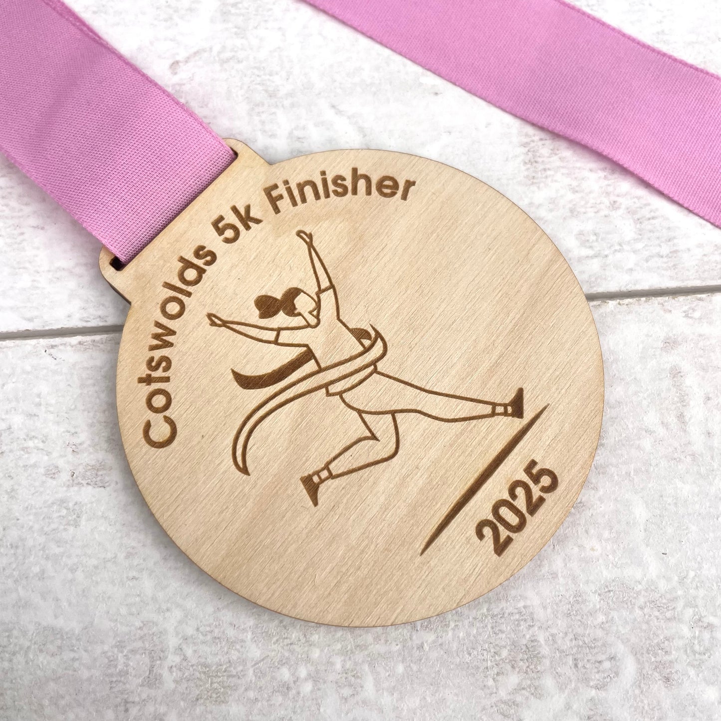 Wooden medal with 'Cotswolds 5k Finisher' text and a pink ribbon on a light wooden surface.
