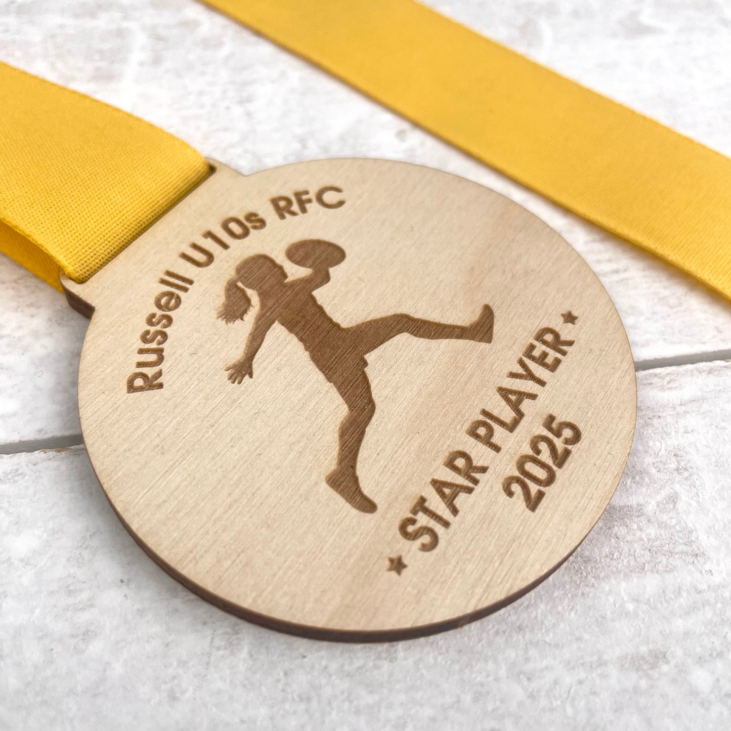 Wooden eco-friendly medal with 'Russell U10s RFC' and 'Star Player 2025' text and silhouette of a rugby player, with a yellow ribbon.