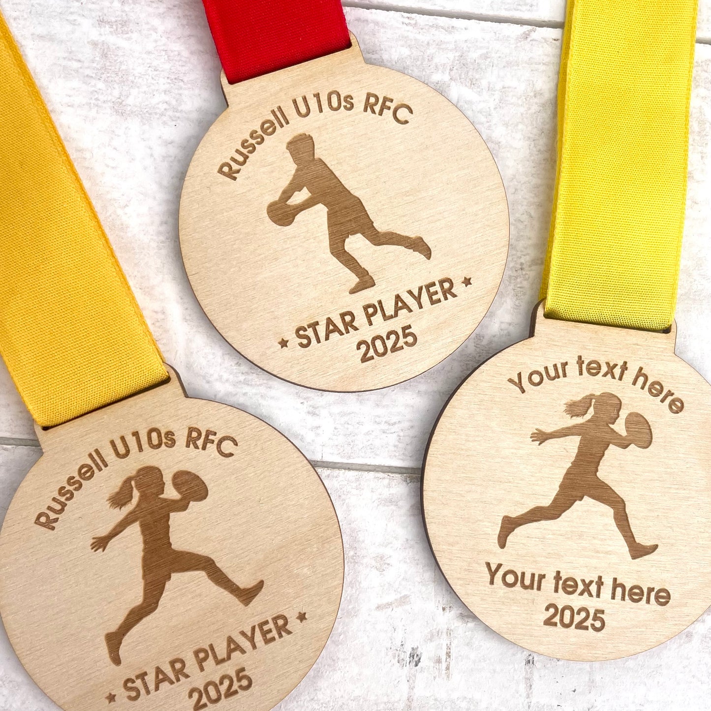 Three eco friendly wooden rugby medals with engraved designs and text with orange, red and yellow ribbons.