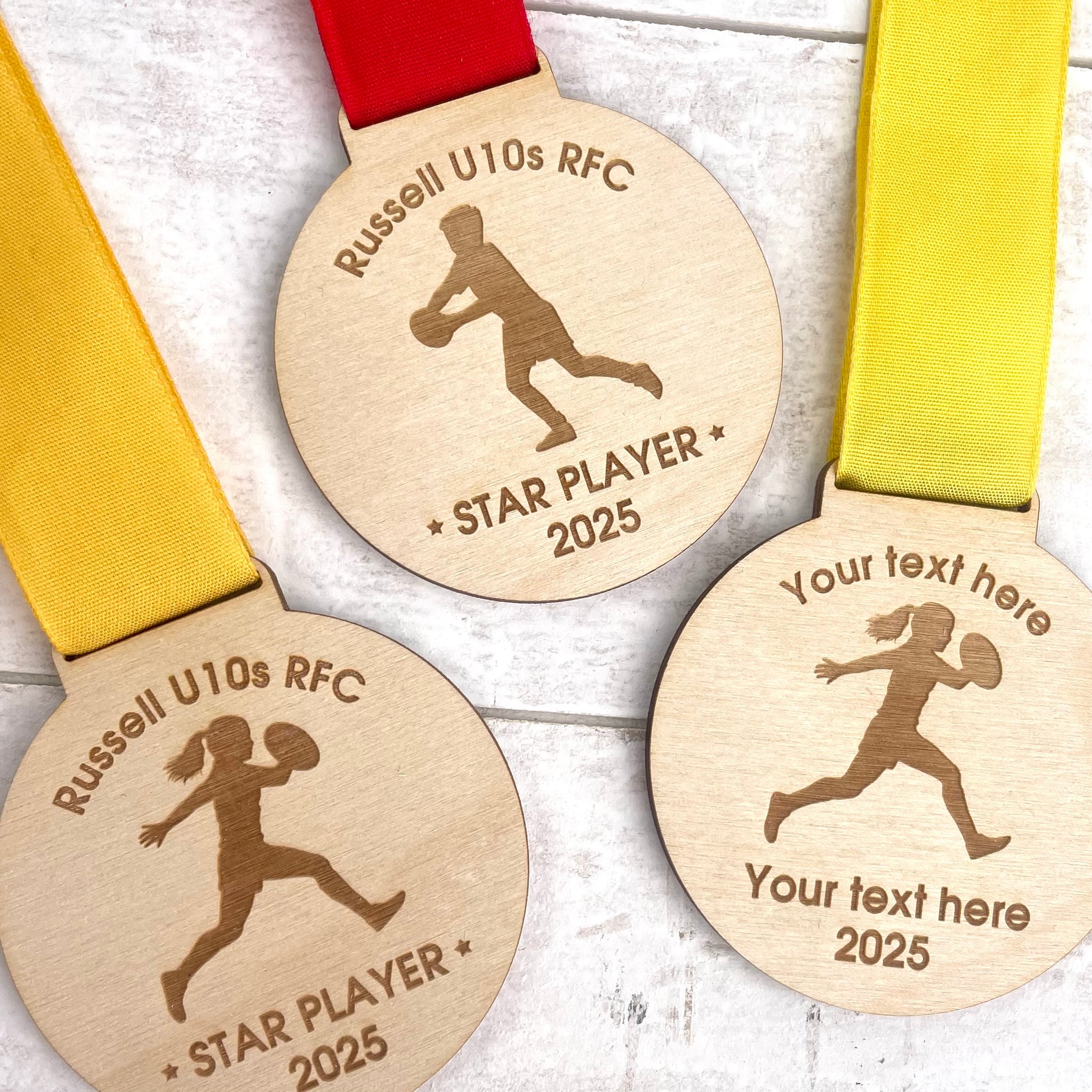 Three eco friendly wooden rugby medals with engraved designs and text with orange, red and yellow ribbons.