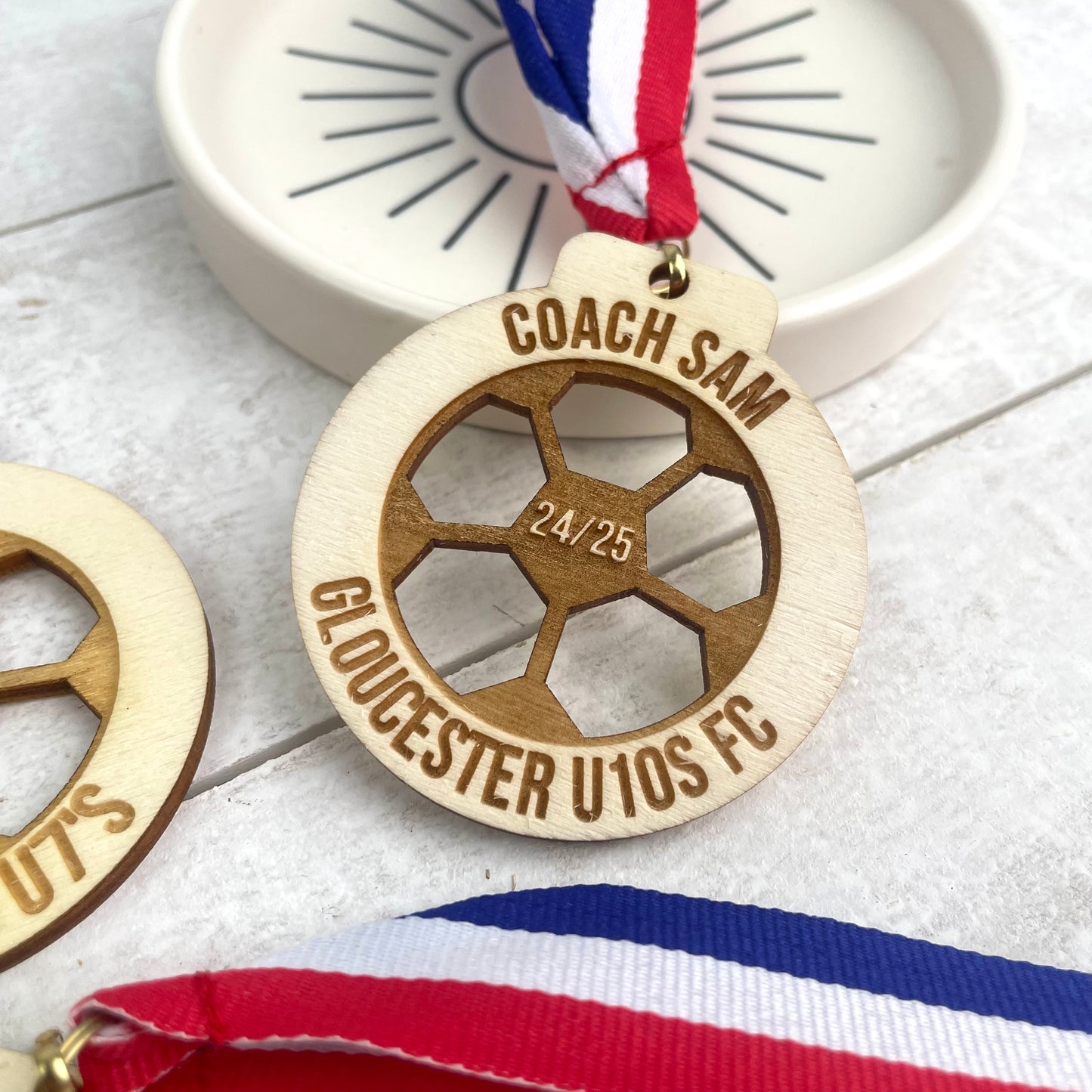 Wooden medal with engraved text 'coach sam' '24/24' 'Gloucester u10s fc' with a striped blue, white and red ribbon.