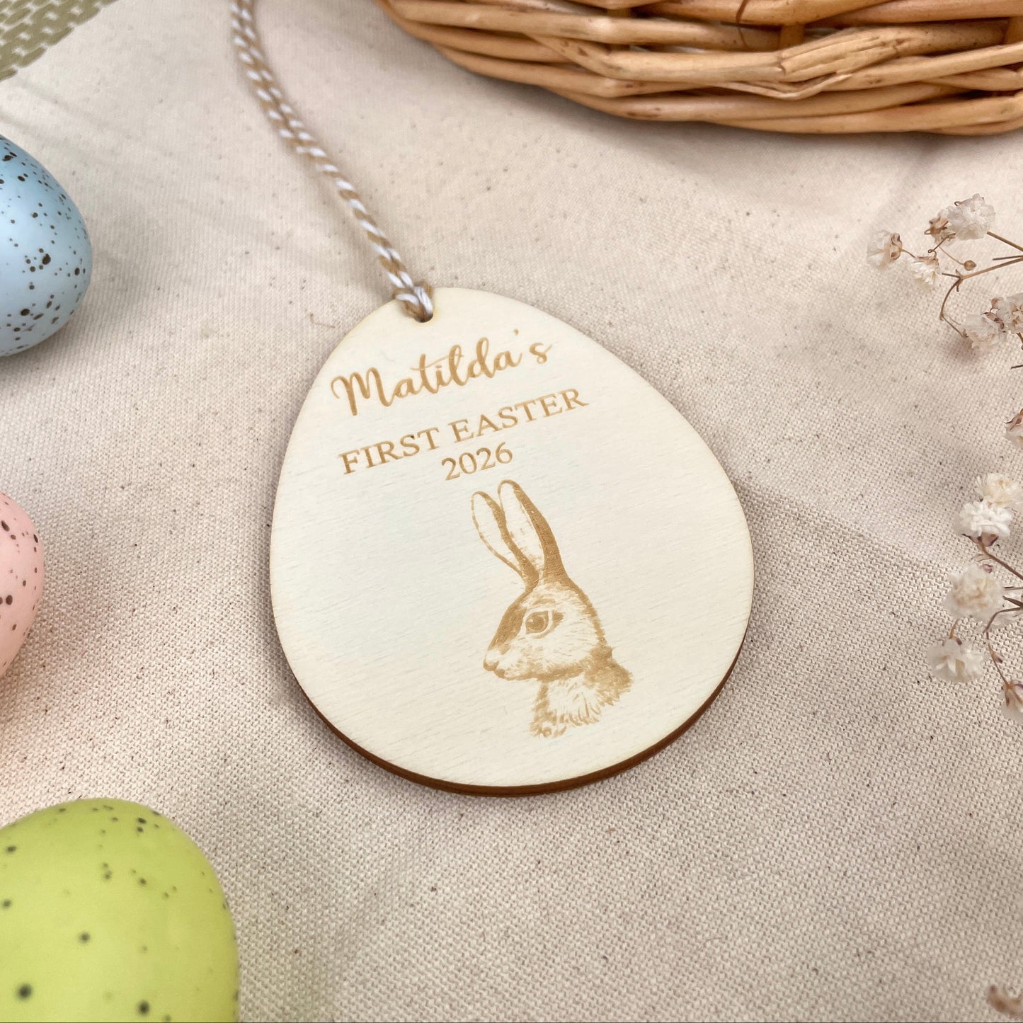 Easter egg shaped personalised wooden decoration with a beige and white string to hang. 'Matilda's First Easter 2026' engraved alongside a hare design. Photo taken on a beige linen with colourful eggs and flowers.