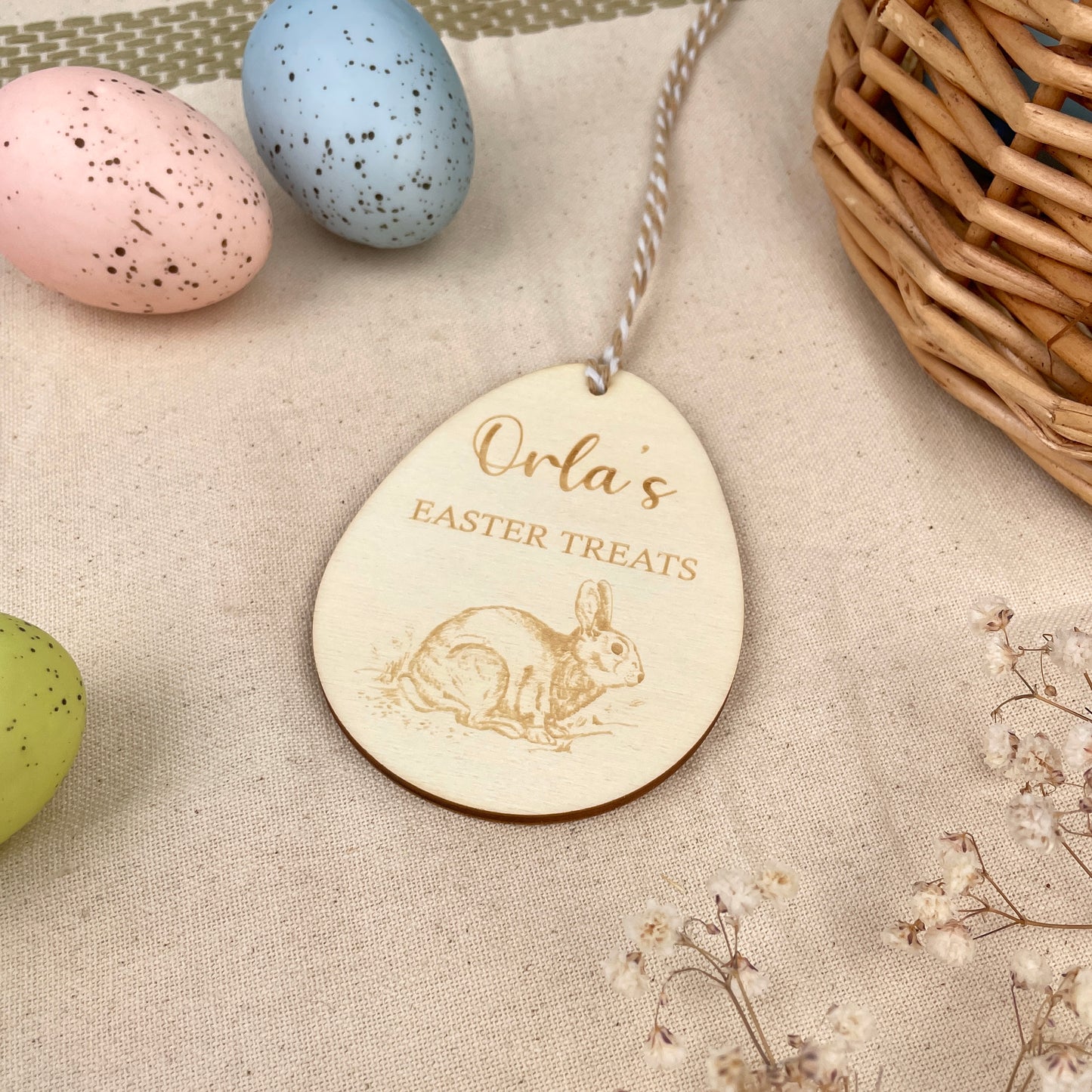 Wooden Easter egg decoration with 'Orla's Easter Treats' engraving and Easter Rabbit illustration, with a beige and white string, on a beige linen with colourful Easter eggs and flowers.