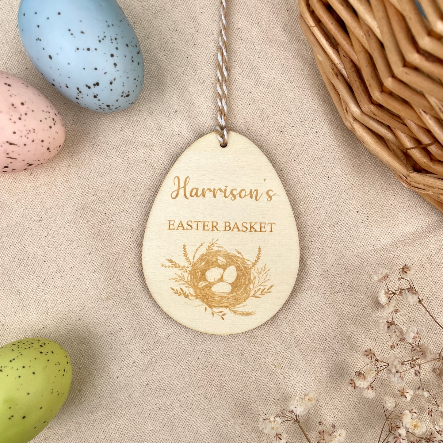 Easter egg shaped personalised wooden decoration with a beige and white string to hang. 'Harrison's Easter Basket' engraved alongside an Easter nest design. Photo taken on a beige linen with colourful eggs and flowers.