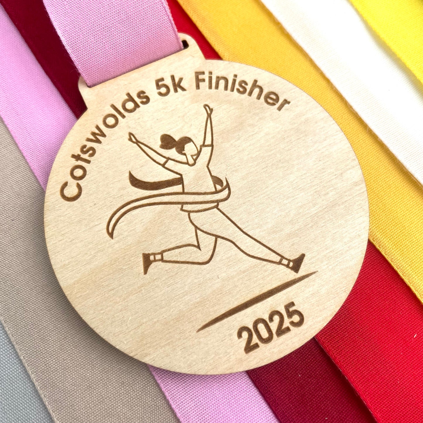 Wooden medal with 'Cotswolds 5k Finisher 2025' engraving and a runner illustration, displayed on colorful ribbons.