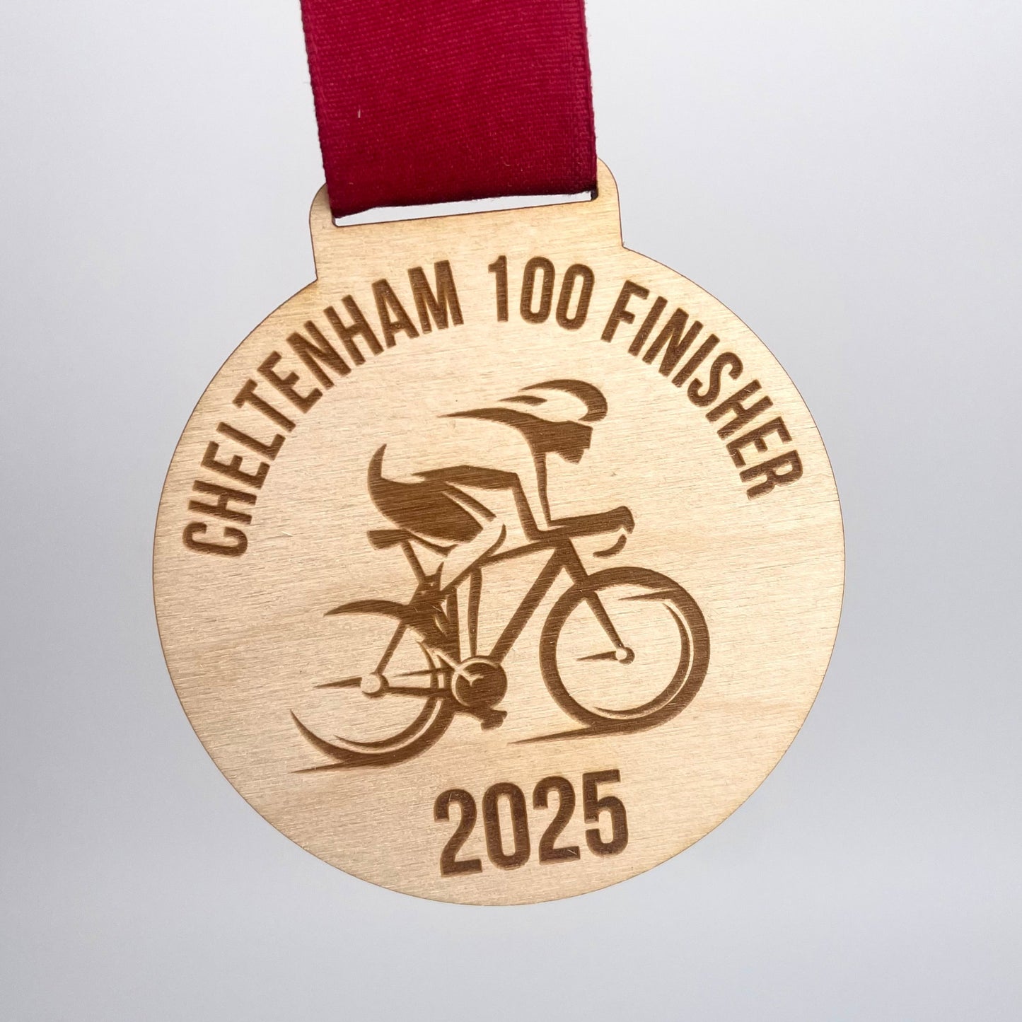 Eco Friendly Wooden medal with 'Cheltenham 100 Finisher 2025' text and a cyclist illustration.