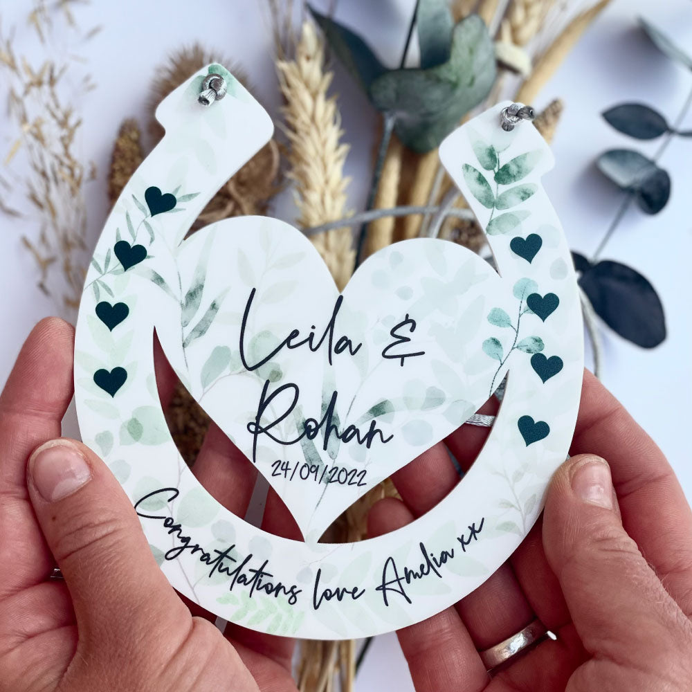 Eucalyptus horseshoe design with engraved names being held up by hands with a floral background front shot