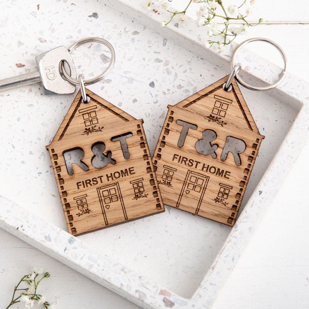 Close up of two engraved new home keyrings with the two initials of the couples cut out, sat on a white stoned background with small white flowers