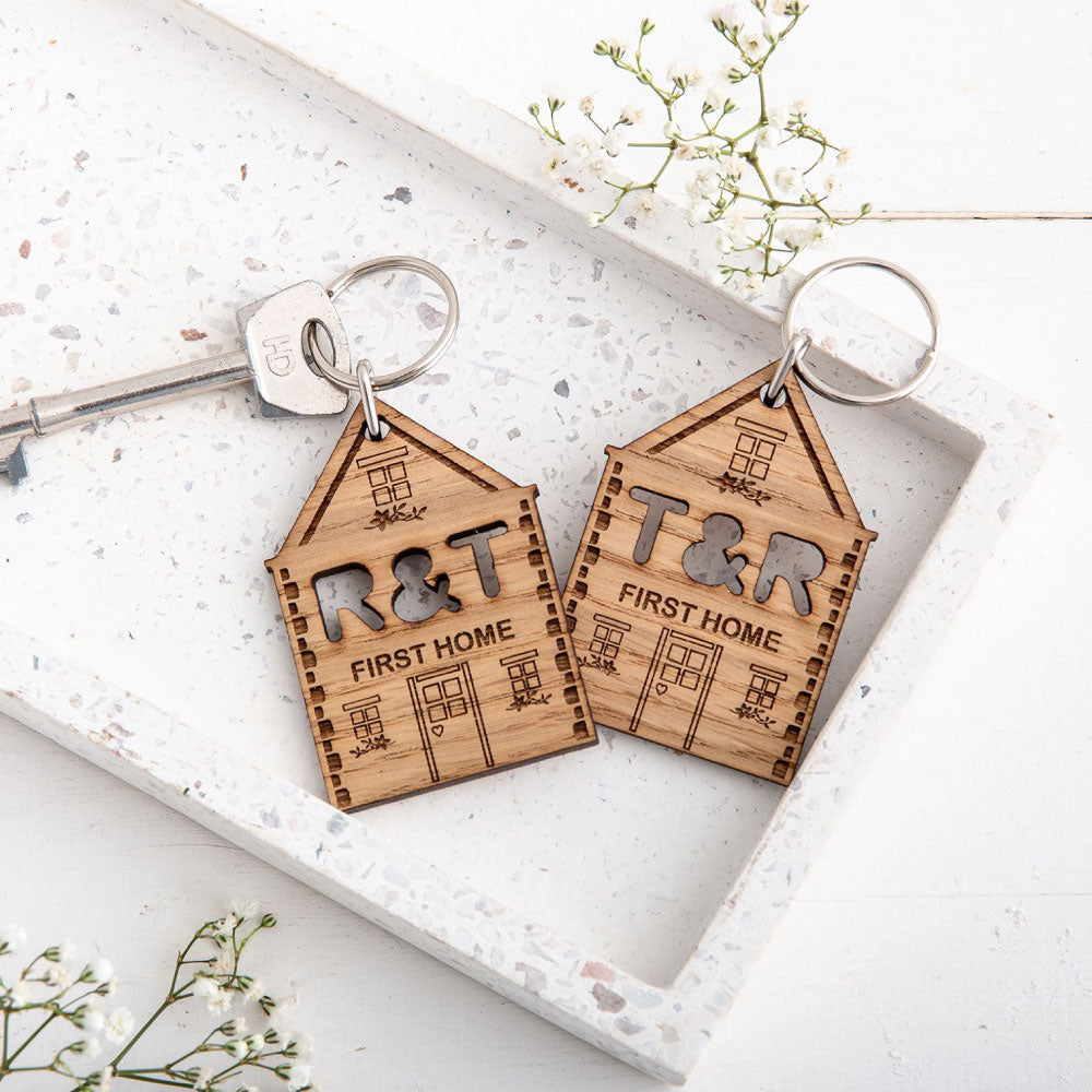 Two engraved new home keyrings with the two initials of the couples cut out, sat on a white stoned background with small white flowers