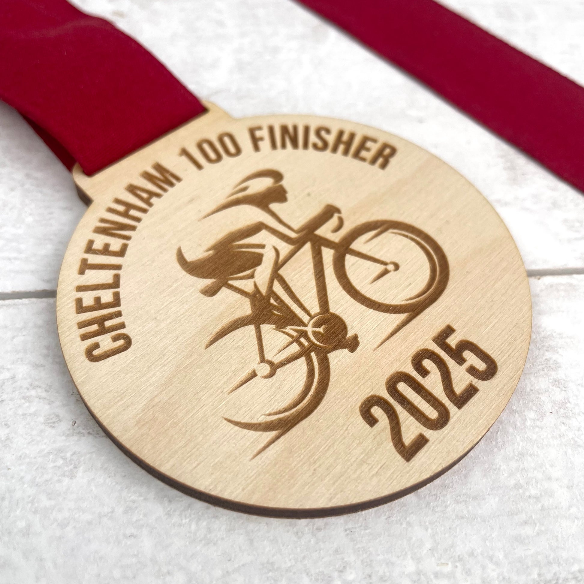 Eco-friendly wooden medal with cyclist illustration and text 'Cheltenham 100 Finisher 2025' on a white background.