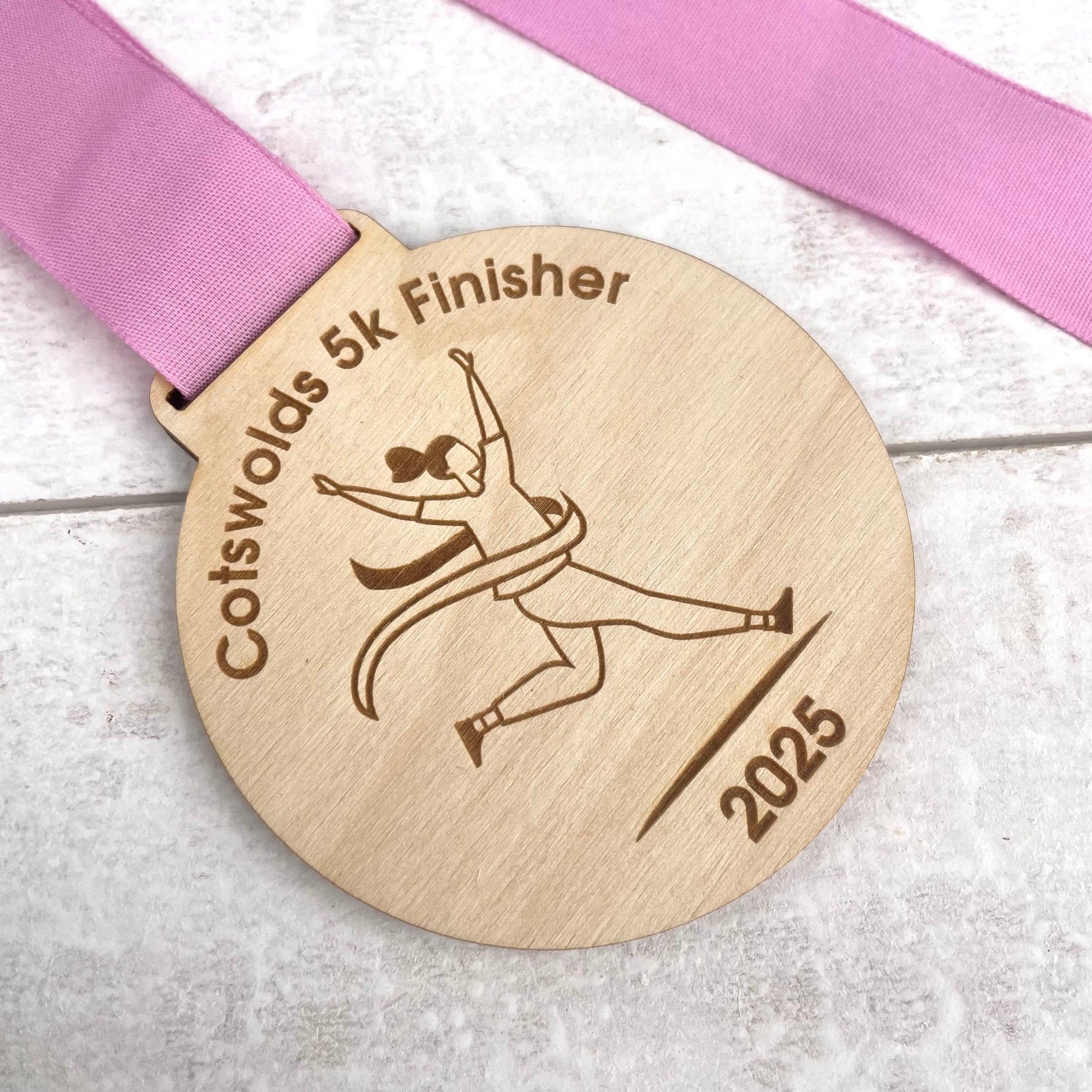 Wooden medal with 'Cotswolds 5k Finisher' text and a pink ribbon on a light wooden surface.