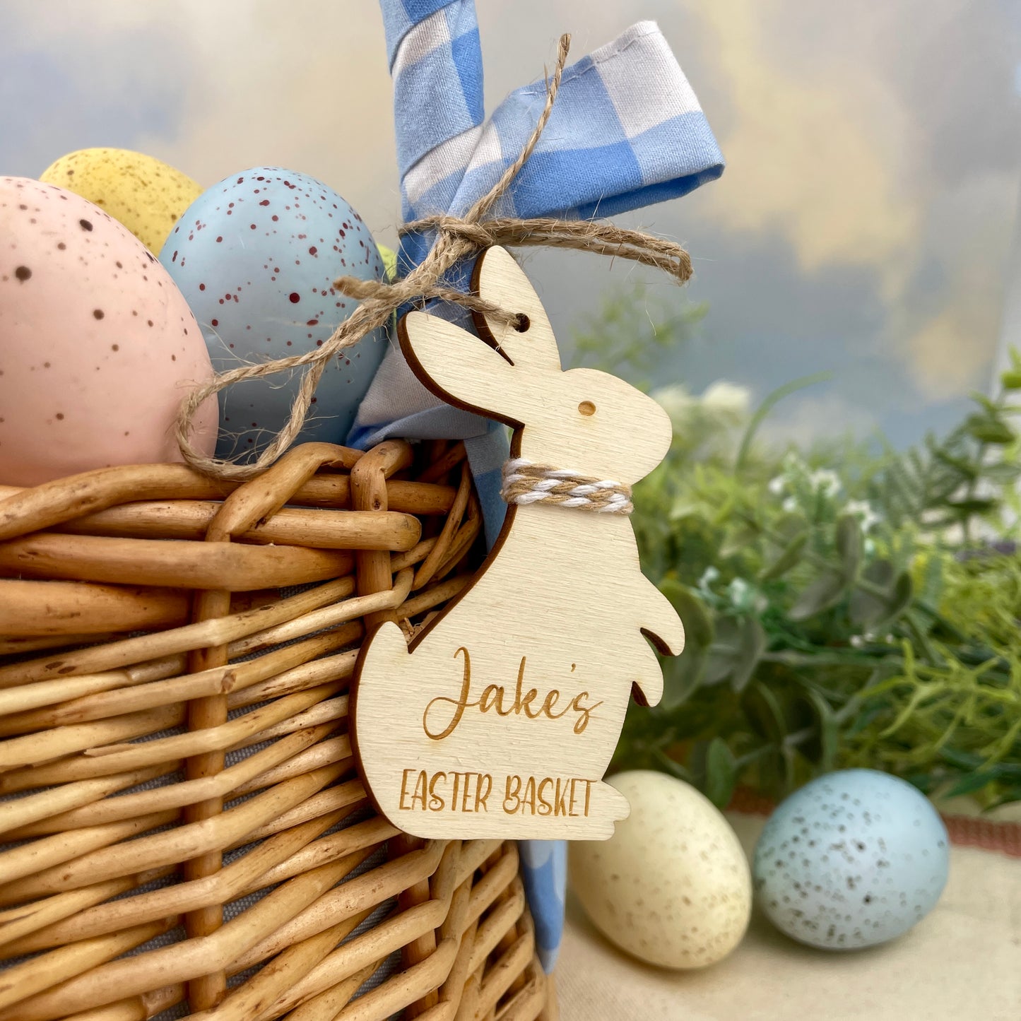Wicker basket with Easter eggs and a wooden personalised Easter bunny tag, with light brown/beige and white string and twine to hang, reading 'Jake's Easter Basket'.