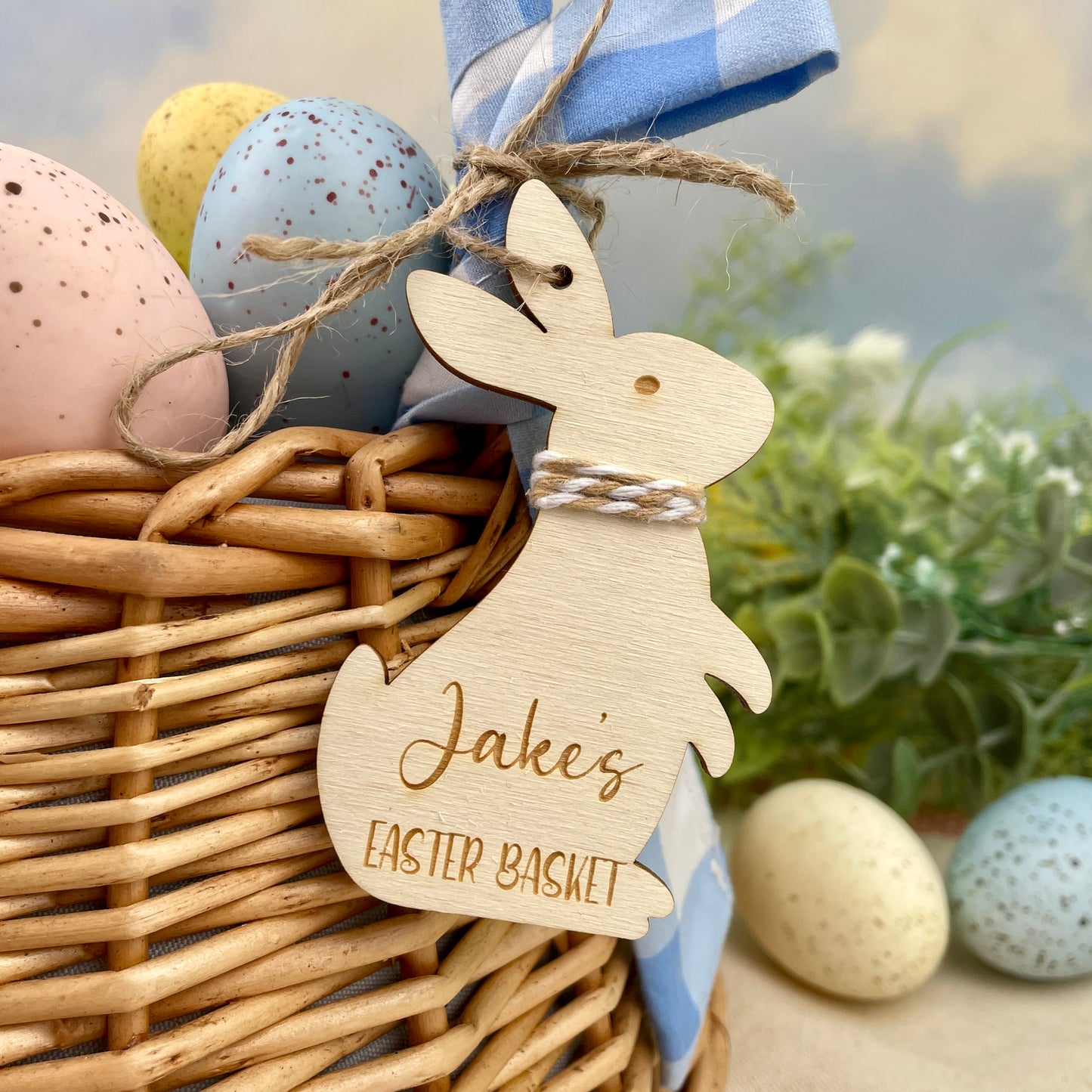 Wicker basket with Easter eggs and a wooden personalised Easter bunny tag, with light brown/beige and white string and twine to hang, reading 'Jake's Easter Basket'.