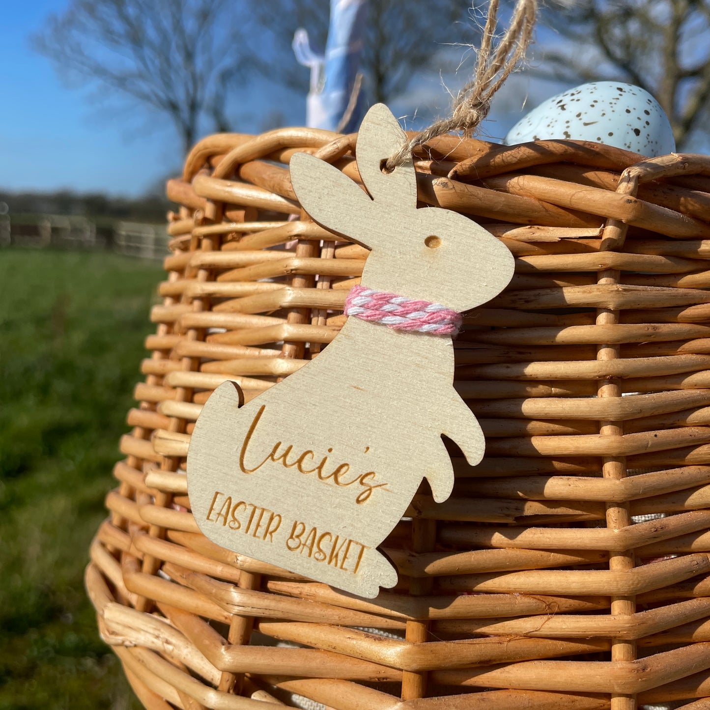 Wicker basket with Easter eggs and a wooden personalised Easter bunny tag, with pink and white string and twine to hang, reading 'Lucie's Easter Basket'.
