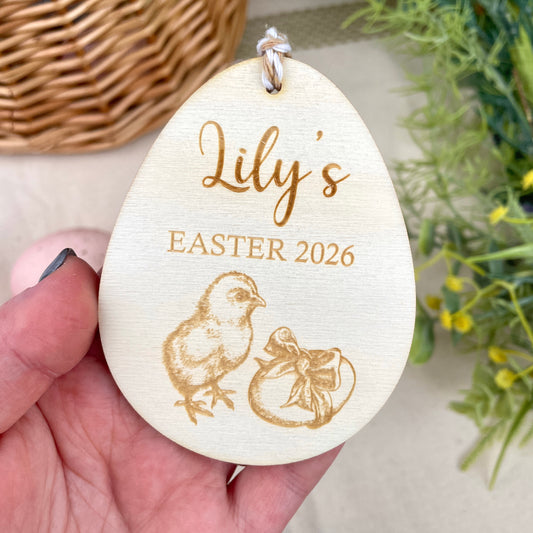 Wooden Easter egg decoration with 'Lily's Easter 2026' engraving and chick illustration, held in a hand.