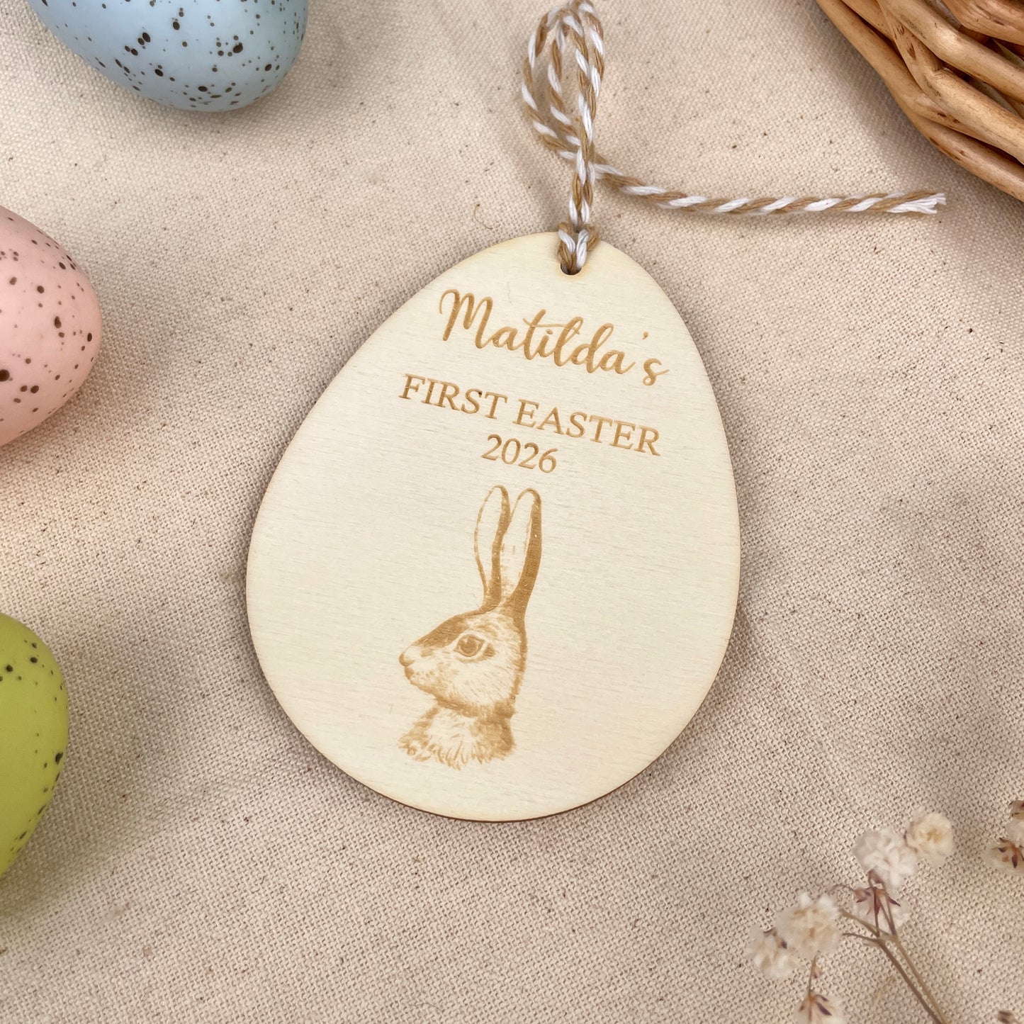 Easter egg shaped personalised wooden decoration with a beige and white string to hang. 'Matilda's First Easter 2026' engraved alongside a hare design. Photo taken on a beige linen with colourful eggs and flowers. 