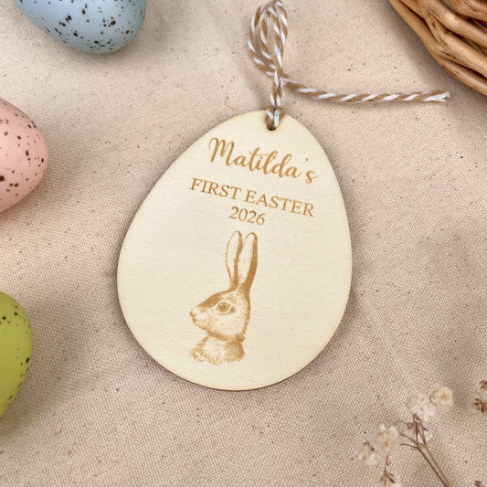 Easter egg shaped personalised wooden decoration with a beige and white string to hang. 'Matilda's First Easter 2026' engraved alongside a hare design. Photo taken on a beige linen with colourful eggs and flowers. 