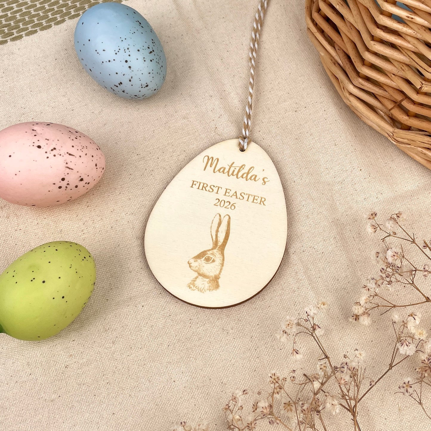 Easter egg shaped personalised wooden decoration with a beige and white string to hang. 'Matilda's First Easter 2026' engraved alongside a hare design. Photo taken on a beige linen with colourful eggs and flowers. 