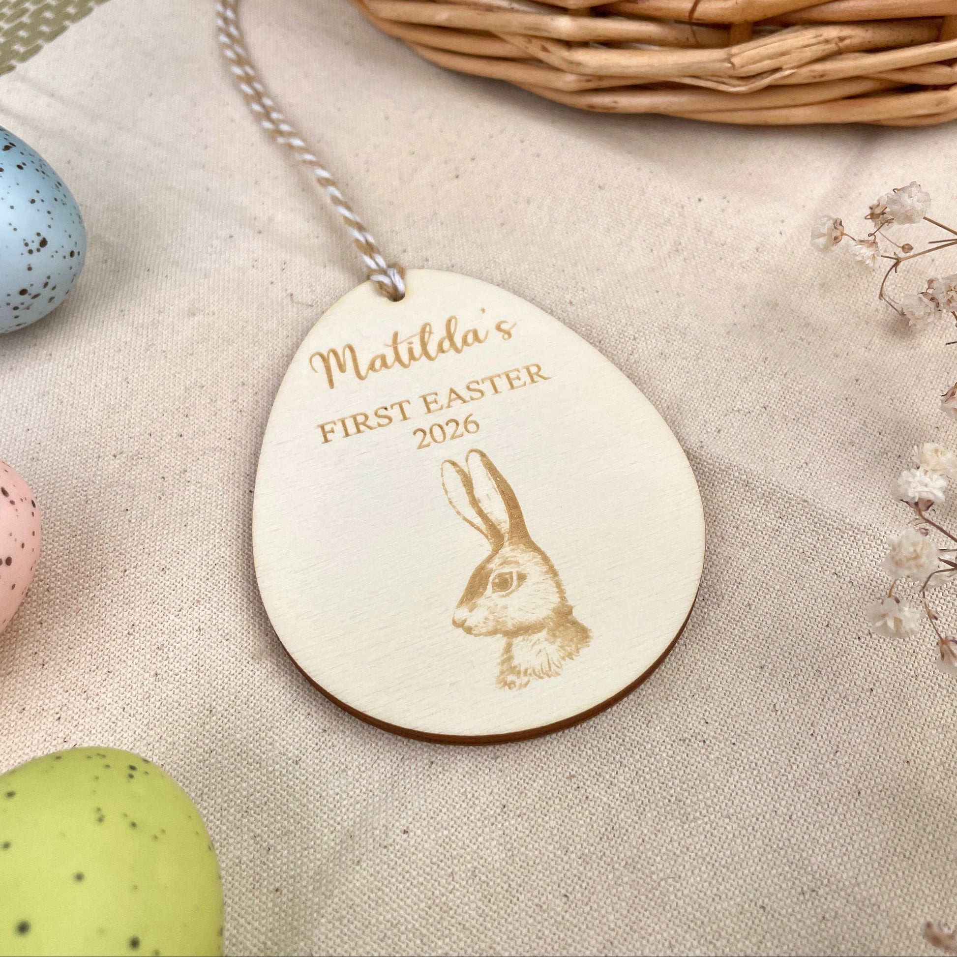 Easter egg shaped personalised wooden decoration with a beige and white string to hang. 'Matilda's First Easter 2026' engraved alongside a hare design. Photo taken on a beige linen with colourful eggs and flowers.  