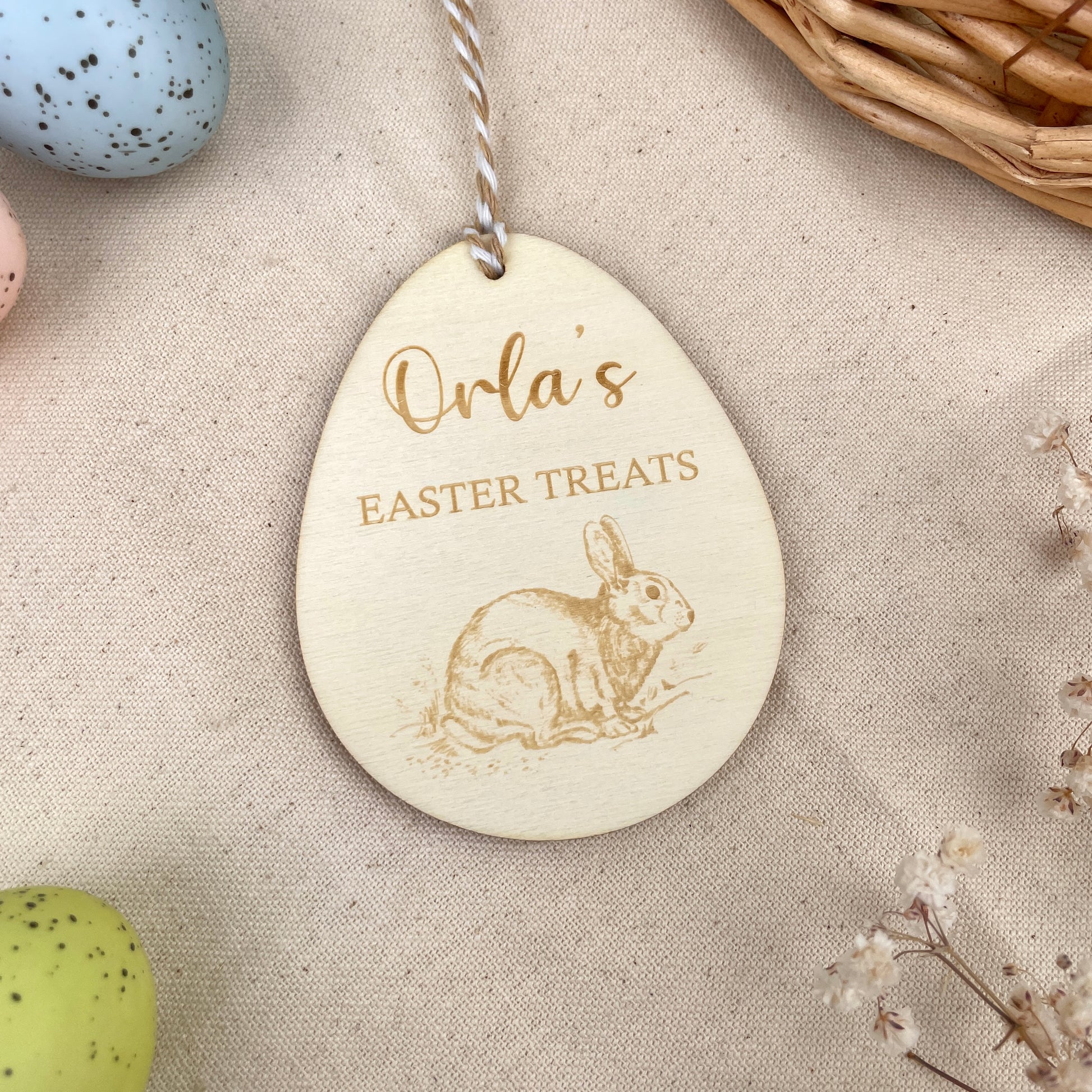 Wooden Easter egg decoration with 'Orla's Easter Treats' engraving and Easter Rabbit illustration, with a beige and white string, on a beige linen with colourful Easter eggs and flowers. 