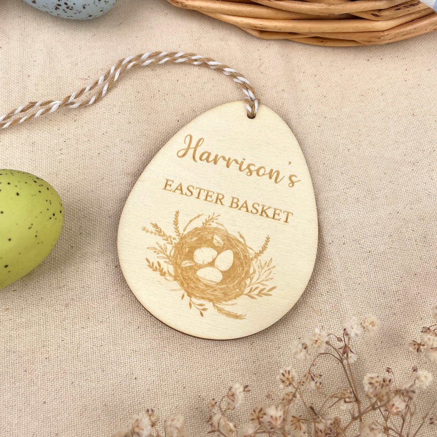 Easter egg shaped personalised wooden decoration with a beige and white string to hang. 'Harrison's Easter Basket' engraved alongside an Easter nest design. Photo taken on a beige linen with colourful eggs and flowers. 