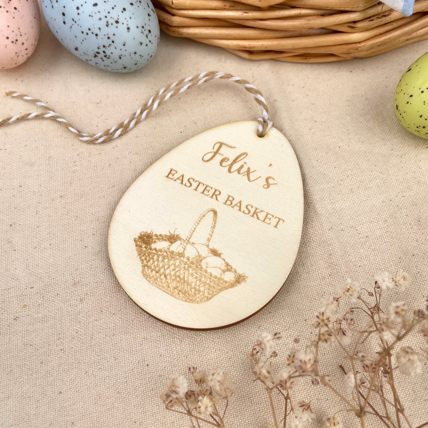 Easter egg shaped personalised wooden decoration with a beige and white string to hang. 'Felix's Easter Basket' engraved alongside an Easter basket design. Photo taken on a beige linen with colourful eggs and flowers. 
