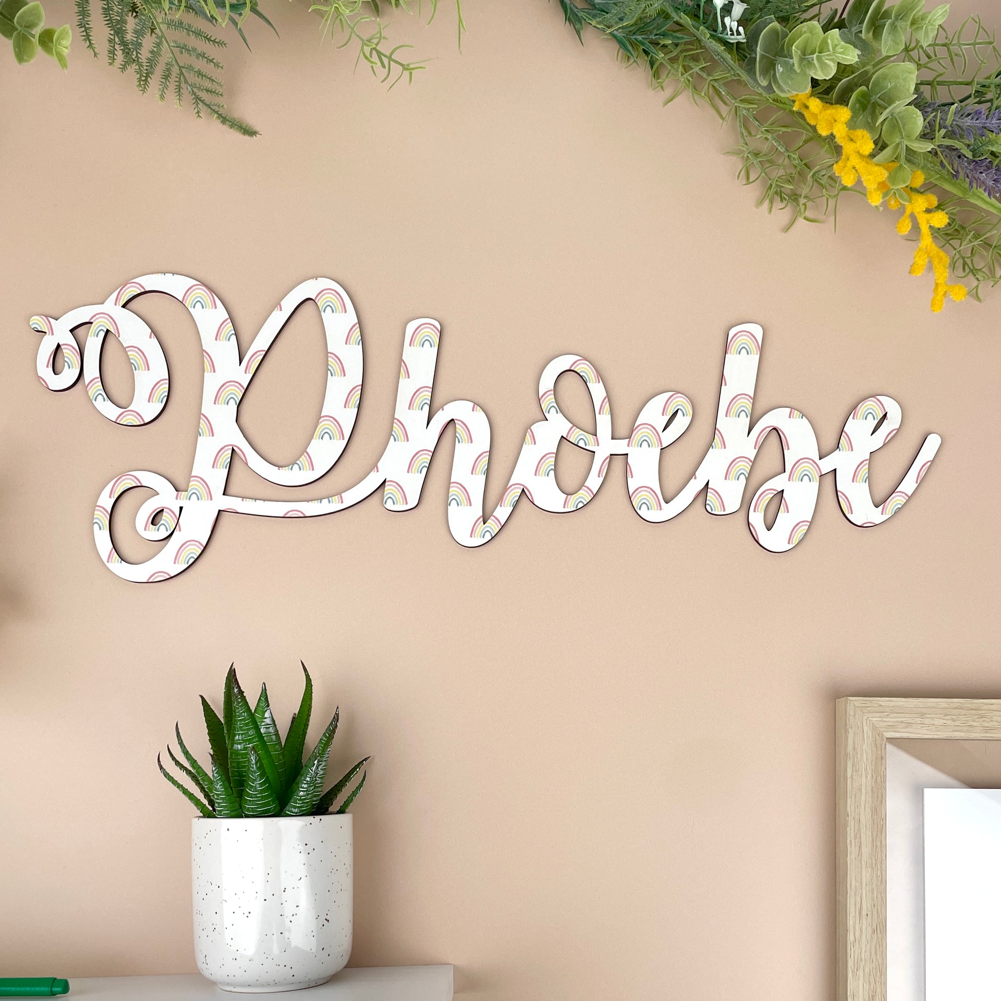 Decorative wooden printed wall sign with 'phoebe' on a beige wall with plants.