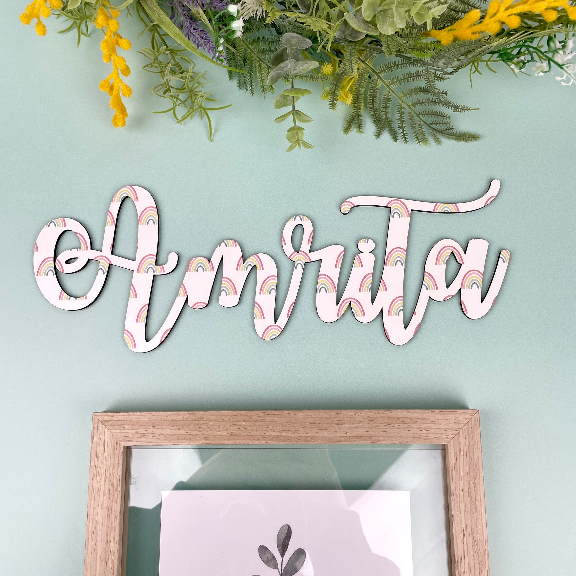 Decorative wall art with 'Amrita' on a light green wall above a framed picture.