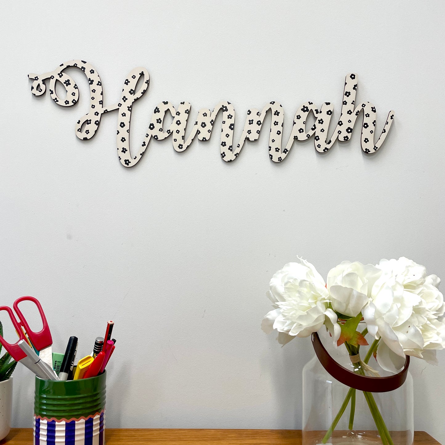Decorative wall art with 'Hannah' on a white wall above a desk with stationery and flowers.