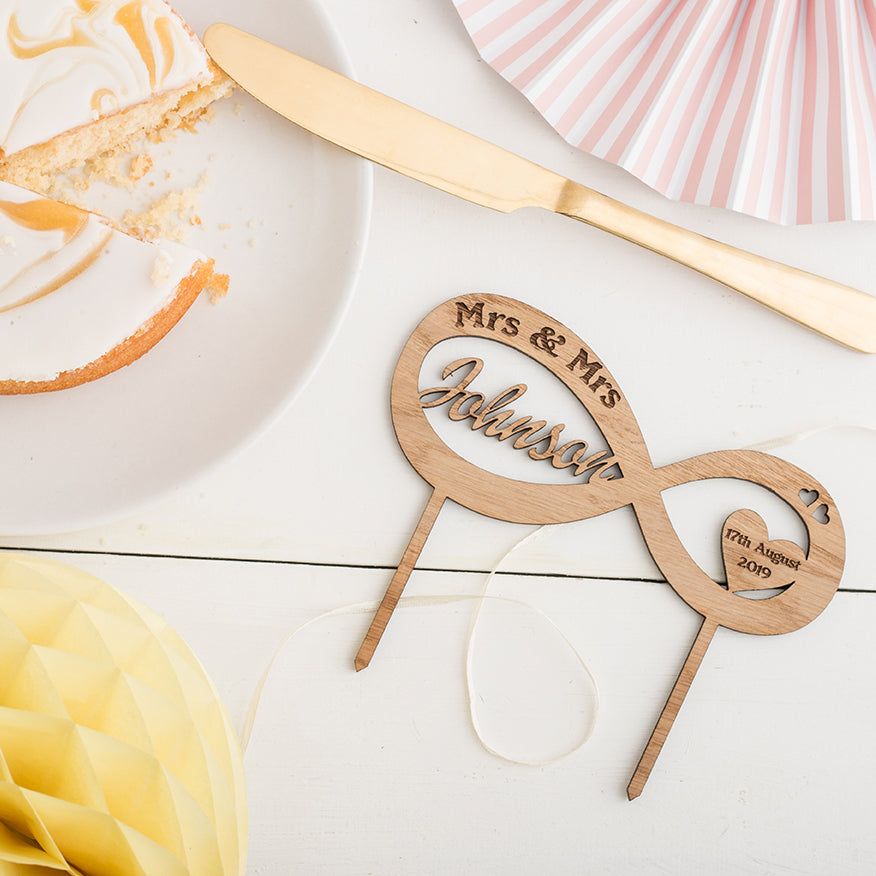 Infinity cake toppper with cut out names and engraved Mr and Mrs wording on the cake topper, sat on a white wooden background surrounded by  party decorations and cake