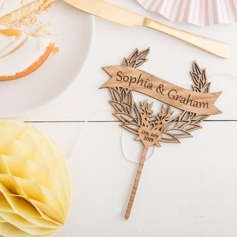 Custom wooden cake topper with personalised names on banner, sat on white wooden background next to a sponge cake and knife