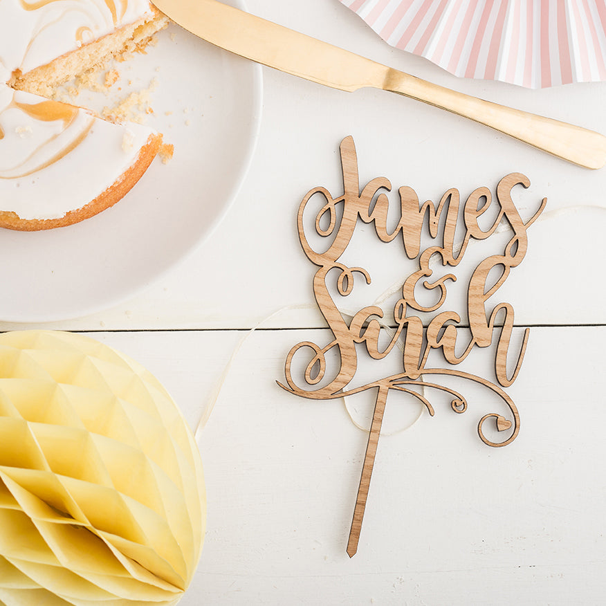 personalised cake topper with the two names james and sarah, on a white background next to cake and party decorations 